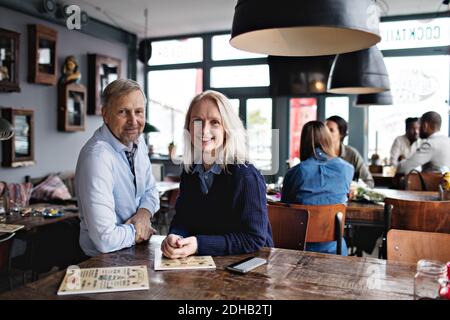 Portrait d'un couple mûr souriant avec menu à une table en bois contre les personnes au restaurant Banque D'Images