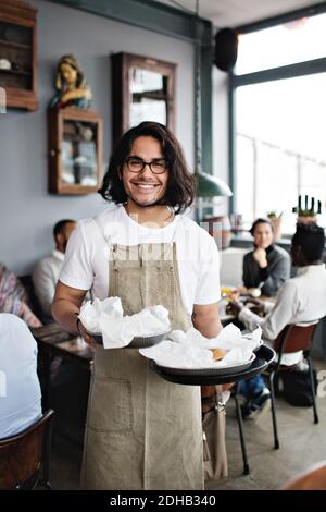 Portrait d'un jeune serveur confiant servant de la nourriture tout en se tenant contre clients au restaurant Banque D'Images