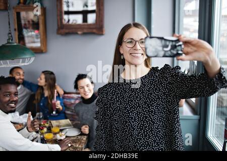 Jeune femme souriante prenant le selfie avec des amis assis au restaurant Banque D'Images