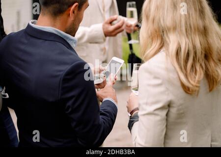 Homme d'affaires montrant un téléphone portable à une femme d'affaires tout en tenant une flûte à champagne Banque D'Images