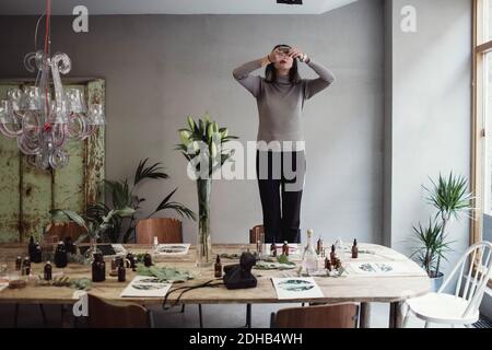 Jeune femme debout sur une chaise, photographiant des flacons de parfum sur une table en atelier Banque D'Images