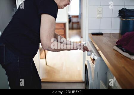 Section médiane d'une femme âgée tenant un téléphone portable tout en utilisant le lavage machine dans la salle de bains à la maison Banque D'Images