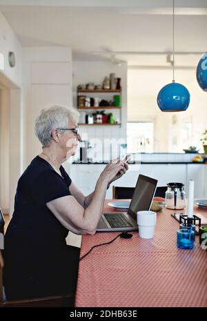 Vue latérale d'une femme âgée en retraite utilisant un smartphone pendant salon avec ordinateur portable à la table à manger Banque D'Images