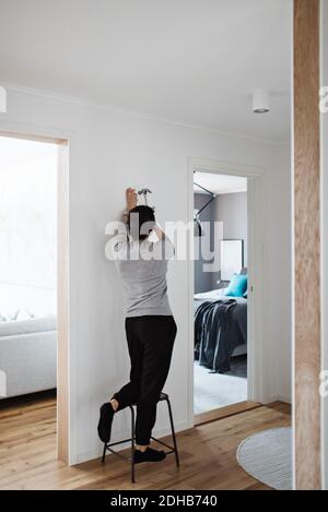 Longueur totale de femme martelant l'ongle sur le mur en se tenant debout sur un tabouret à la maison Banque D'Images