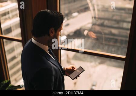 Vue en grand angle de l'avocat masculin à l'aide d'un smartphone par fenêtre au bureau Banque D'Images