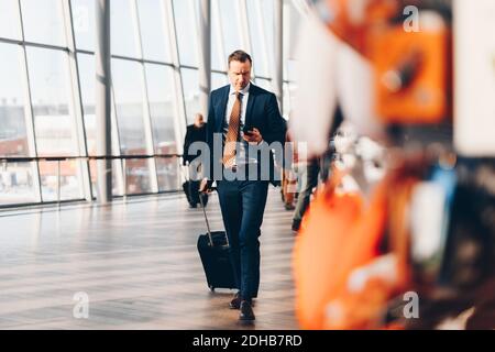 Pleine longueur d'homme d'affaires mature avec bagages à l'aide d'un téléphone portable en marchant dans le terminal de l'aéroport Banque D'Images