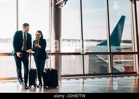 Couple d'affaires pleine longueur avec bagages à l'aide d'un téléphone portable à l'aéroport Banque D'Images