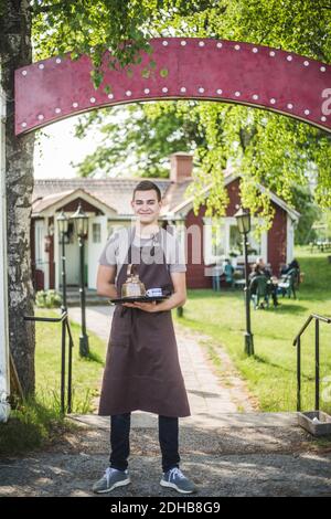 Portrait complet d'un jeune serveur souriant tenant un plateau de service en vous tenant à l'entrée du café extérieur Banque D'Images