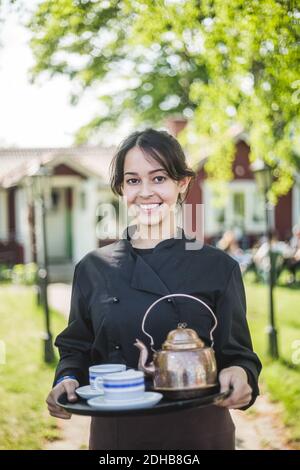Portrait d'une serveuse jeune femme confiante souriant tout en tenant le service plateau contre le restaurant Banque D'Images