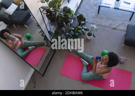 Vue aérienne d'une femme afro-américaine assise sur un tapis d'exercice et l'eau potable. Auto-isolation fitness à la maison pendant la pandémie du coronavirus Covid 19. Banque D'Images