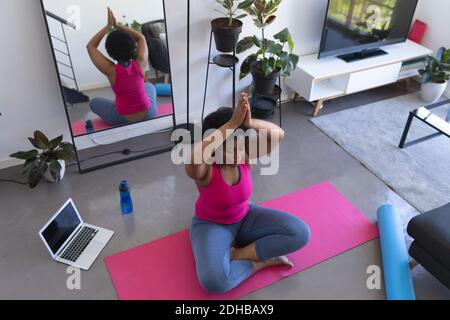 Femme afro-américaine faisant de la méditation de yoga assis sur un tapis portant des vêtements de sport. ordinateur portable en arrière-plan. Auto-isolation fitness bien-être technol Banque D'Images