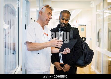 Un médecin senior discute avec un jeune homme au sujet d'une tablette numérique pendant contrôle de routine dans le couloir Banque D'Images