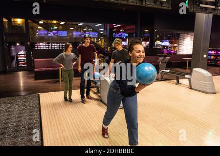 Adolescente souriante tenant le ballon contre des amis debout sur éclairé parquet au bowling Banque D'Images