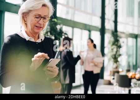 Entrepreneur principal utilisant le téléphone mobile pendant que ses collègues discutent en arrière-plan Banque D'Images