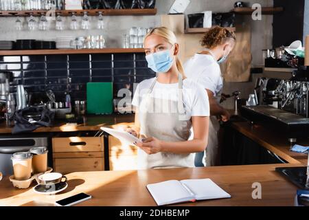 un serveur blond dans un masque médical tenant une tablette numérique près d'un ordinateur portable, un café à emporter et un barista travaillant en arrière-plan Banque D'Images