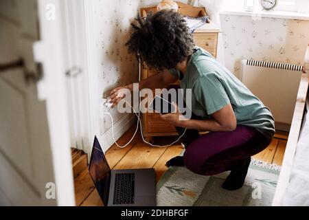 Vue latérale d'une jeune femme qui branche le chargeur de téléphone portable prise électrique à la maison vue par la porte Banque D'Images