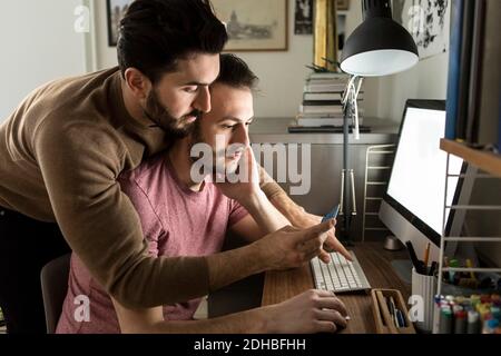 Gay couple regardant la carte de crédit tout en faisant des achats en ligne sur ordinateur à la maison Banque D'Images
