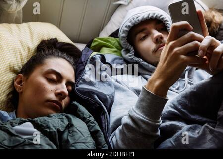 Vue en grand angle d'un jeune homme à l'aide d'un téléphone portable pendant couché par un ami sur le lit à la maison Banque D'Images