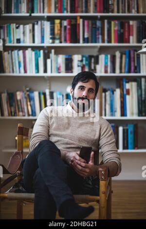 Portrait d'un jeune homme tenant un smartphone assis contre lui bibliothèque Banque D'Images