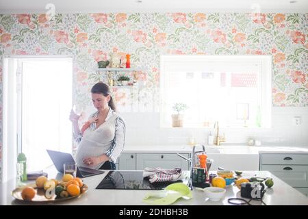 Femme blogueuse utilisant un ordinateur portable tout en portant la fille dans la cuisine à accueil Banque D'Images