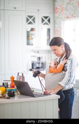 Vue latérale de la nourriture blogger avec l'appareil photo utilisant l'ordinateur portable pendant porter bébé fille dans la cuisine Banque D'Images