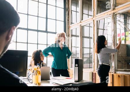 Les collègues regardent une femme d'affaires qui colle des notes adhésives sur un mur en verre dans la salle du conseil pendant la réunion Banque D'Images