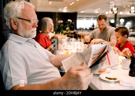 Homme senior lisant le journal pendant qu'il est assis en famille dans le restaurant pendant petit déjeuner Banque D'Images