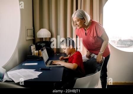 Grand-mère regardant le petit-fils à l'aide d'un ordinateur portable assis près d'une table dans la chambre d'hôtel Banque D'Images