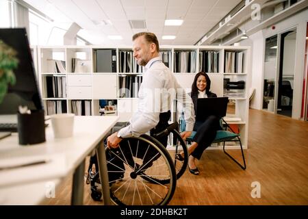 Homme handicapé professionnel assis sur un fauteuil roulant tout en souriant femme d'affaires utilisant ordinateur portable sur le lieu de travail Banque D'Images