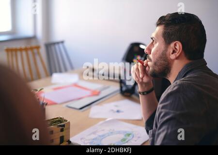 Vue latérale d'un ingénieur en train de penser tout en étant assis avec le schéma à la table dans le bureau Banque D'Images