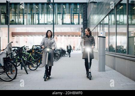 Longueur totale de collègues féminins confiants à la conduite d'un scooter électrique sur le sentier en bâtiment dans la ville Banque D'Images
