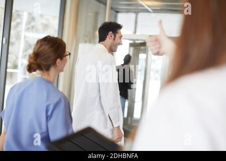 Image rognée de l'infirmière féminine montrant les pouces jusqu'à jeune médecin marchant avec un collègue dans le hall de l'hôpital Banque D'Images