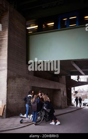 Des amis adolescents, hommes et femmes, avec des scooters de poussée et des vélos sur le chemin de pied sous le pont Banque D'Images