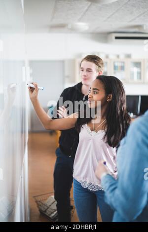 Une jeune fille confiante écrit sur un tableau blanc tout en se tenant au milieu d'un professeur et ami en classe Banque D'Images