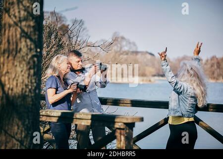 Homme et femme senior photographiant un ami à travers un appareil photo tout en étant debout par lac Banque D'Images