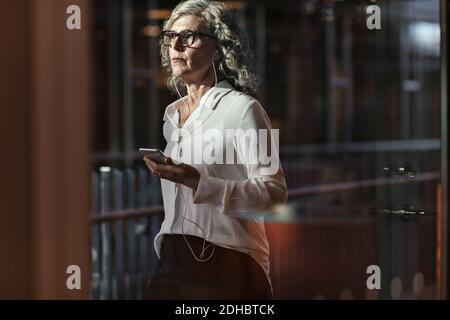 Femme d'affaires sénior qui porte des écouteurs tout en utilisant un téléphone portable à bureau Banque D'Images