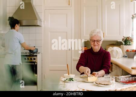 Souriante femme âgée à la retraite prenant le petit-déjeuner tandis que la femme soignante prépare nourriture sur cuisinière dans la cuisine à la maison de repos Banque D'Images