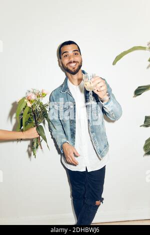 Main courte de femme donnant des fleurs au jeune homme souriant tenir avec une boisson tout en se tenant contre le mur blanc à la maison Banque D'Images
