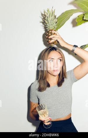 Jeune femme joueur portant de l'ananas sur la tête tout en étant debout avec buvez contre le mur blanc à la maison Banque D'Images