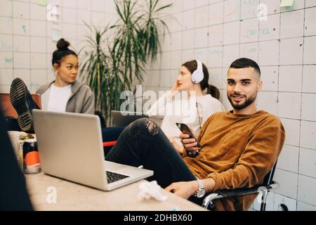Portrait d'un jeune homme d'affaires souriant tenant un smartphone assis avec des collègues féminins dans un petit bureau Banque D'Images