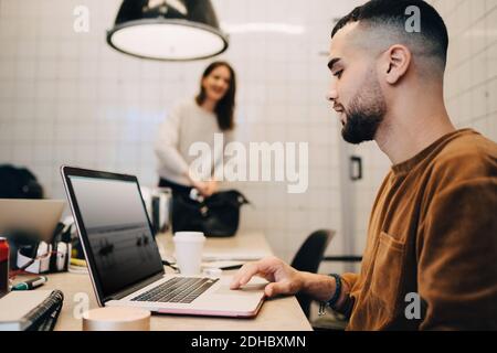Vue latérale d'un jeune hacker mâle utilisant un ordinateur portable alors qu'une femme collègue travaillant dans un petit bureau Banque D'Images