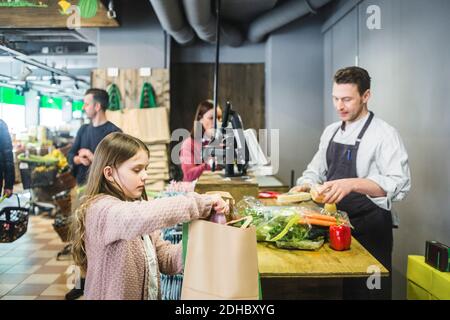 Une petite fille mignonne insère des articles d'épicerie dans un sac en papier au comptoir des caisses Banque D'Images
