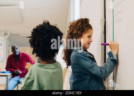 Garçon debout près d'une fille écrivant sur un tableau blanc en classe Banque D'Images