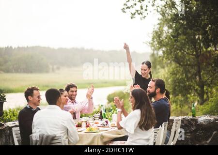 Femme adulte de taille moyenne debout avec le bras levé pendant que les amis applaudissent à la table pendant le dîner dans l'arrière-cour Banque D'Images