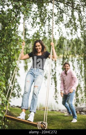 Femme gaie balançant sur corde balançoire alors que l'ami mâle debout au bord du lac Banque D'Images