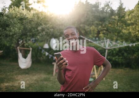 Portrait d'un garçon souriant tenant un téléphone portable debout arrière-cour pendant la fête de jardin Banque D'Images