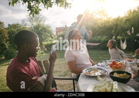 Garçon photographiant une famille joyeuse en dégustant un déjeuner à table pendant le jardin fête Banque D'Images