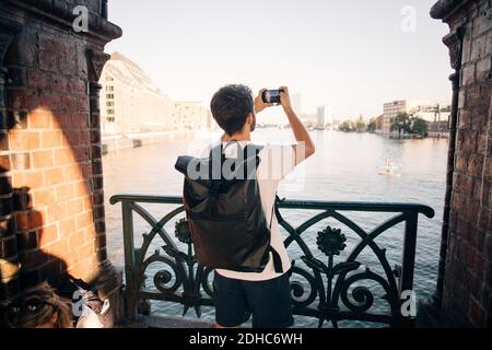 Vue arrière d'un jeune homme qui photographie la rivière à l'aide d'un smartphone en ville Banque D'Images