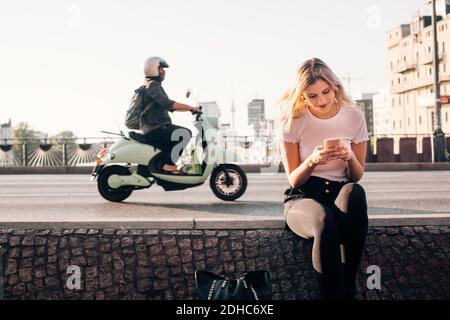 Jeune femme souriante utilisant un smartphone tout en restant assise mur dans la rue en ville Banque D'Images