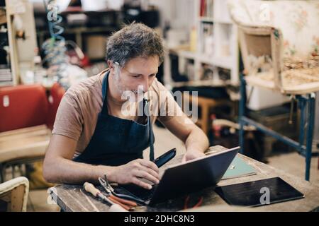 Homme sellerie utilisant un ordinateur portable assis à l'établi à l'intérieur atelier Banque D'Images